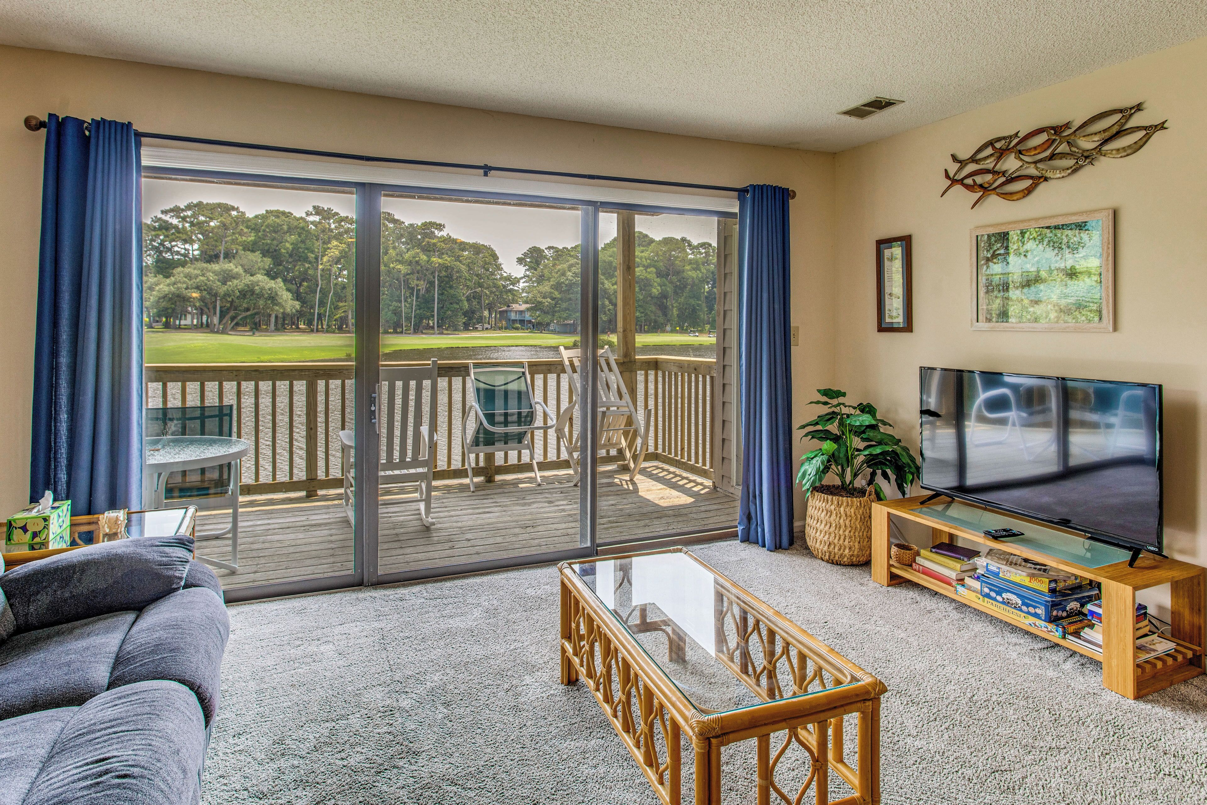 A living room with a large sliding glass door leading to a deck, a flat-screen TV, a coffee table, and a bookshelf.