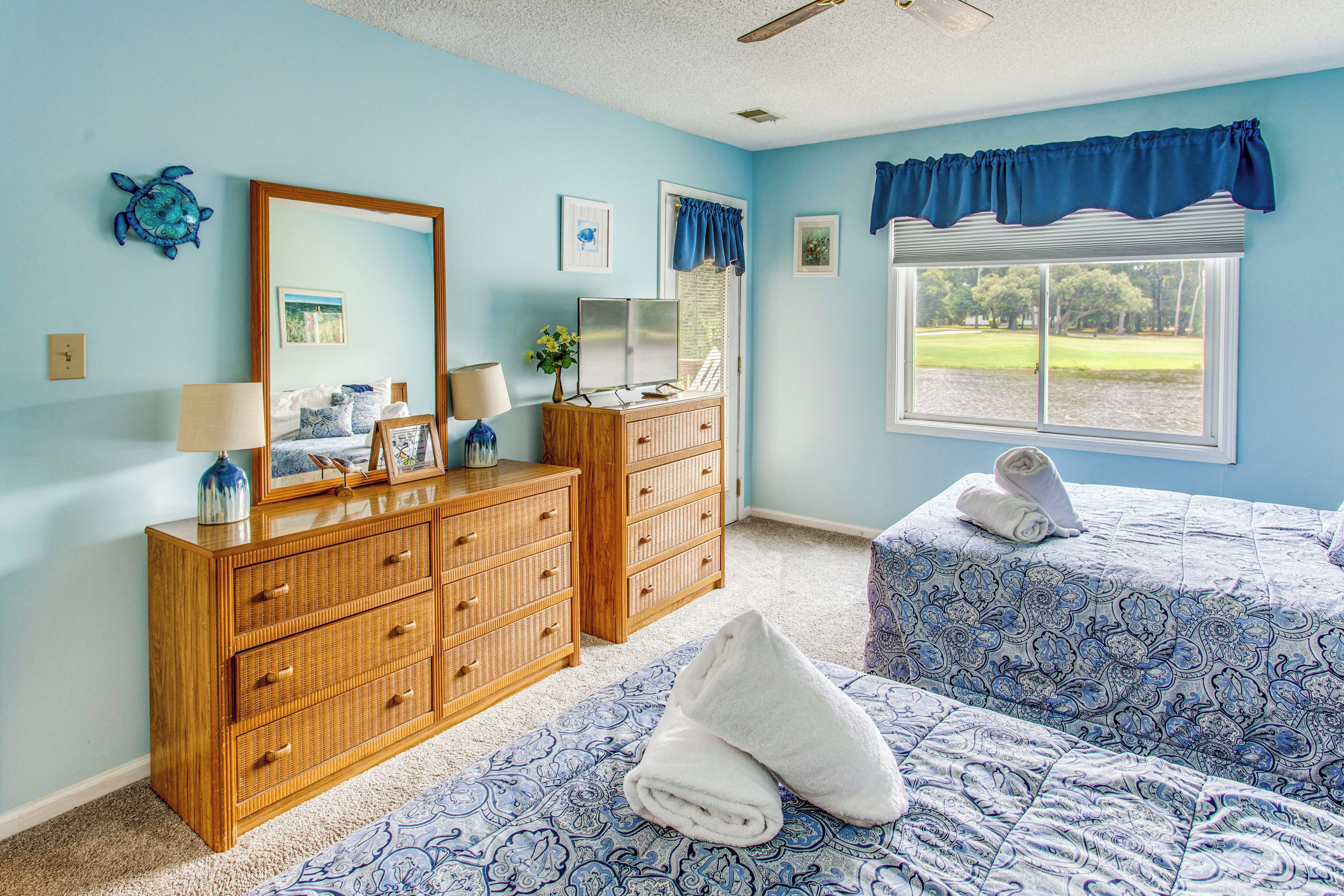 A bedroom with two beds, a dresser, a mirror, and a window with a view of a park.