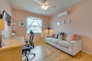 Ein Zimmer mit Schreibtisch, Stuhl, Sofa, Deckenventilator, einem Fenster mit Jalousien und Wandaircondition.