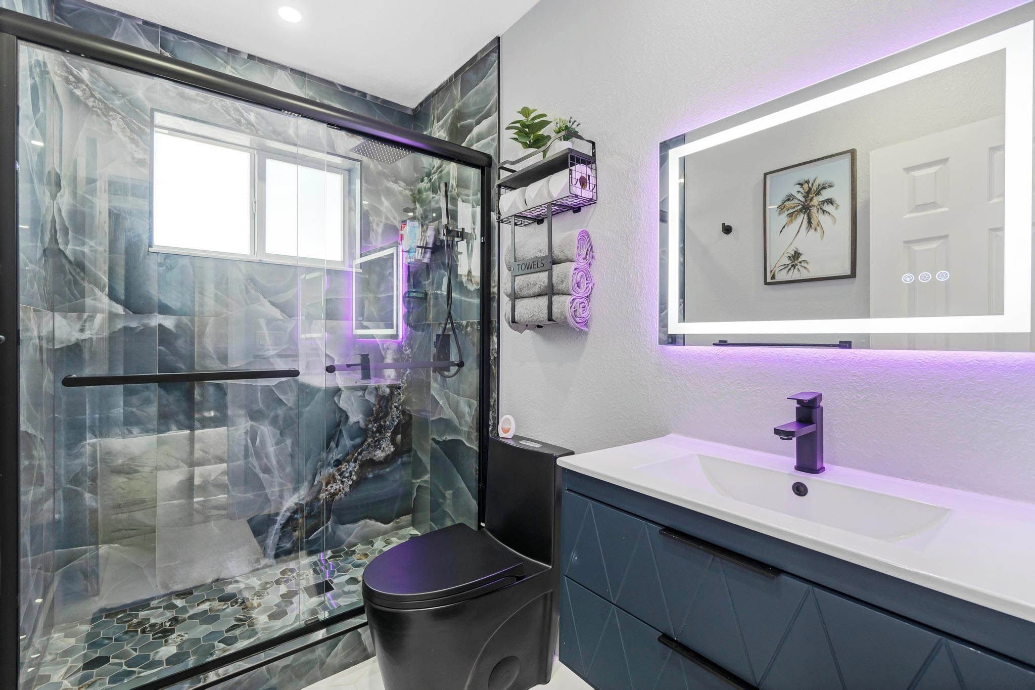 Un baño moderno con cabina de ducha de vidrio, un lavabo blanco con grifo morado y un cuadro enmarcado sobre el espejo.