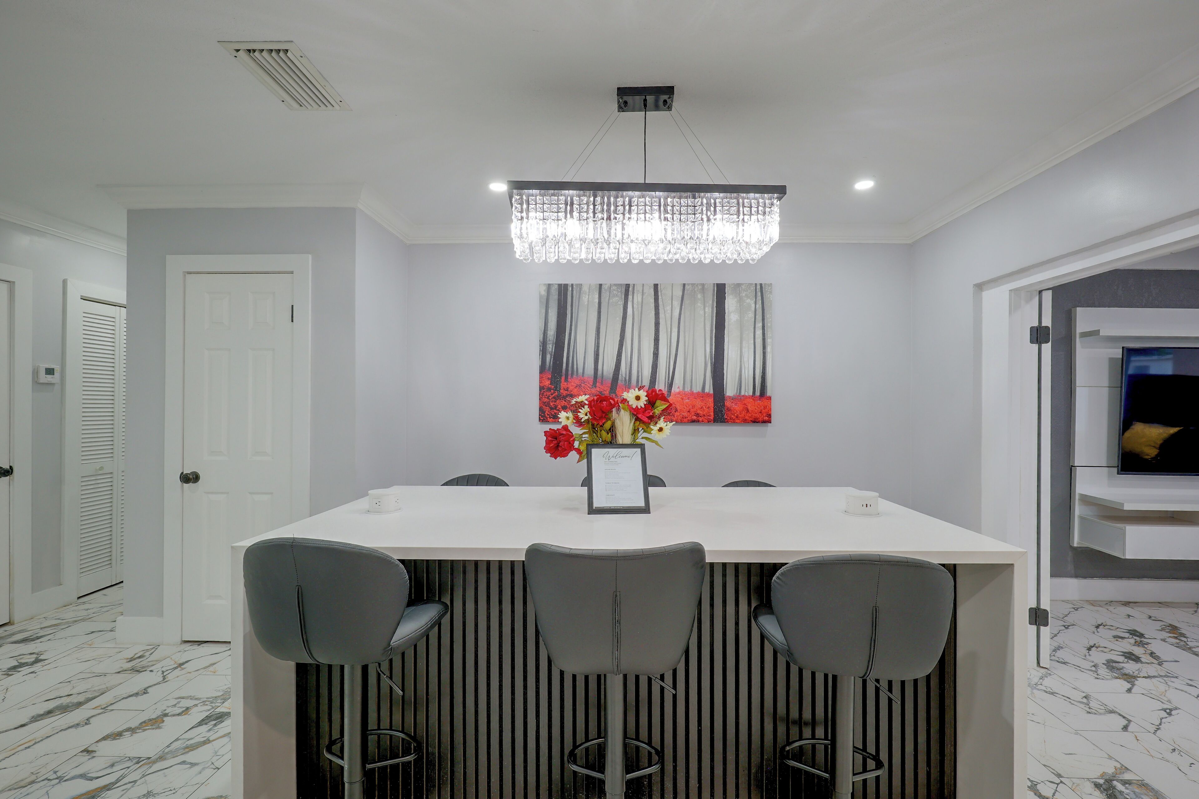 A modern dining area with a white table, bar stools, a chandelier, and a wall-mounted TV.