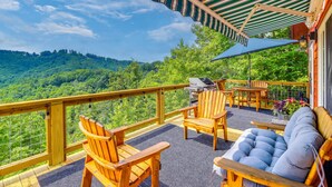 Eine Holzterrasse mit Gartenmöbeln, einem Grill und Blick auf einen bewaldeten Hang.