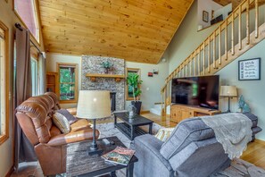 Ein geräumiges Wohnzimmer mit Kamin aus Stein, Ledersofas, einem Flachbildfernseher und einer Holztreppe.