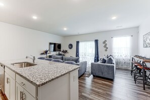 A modern kitchen with granite countertops and a stainless steel sink, a living area with grey sofas and a coffee table, and a dining area with a wooden table and bar stools.
