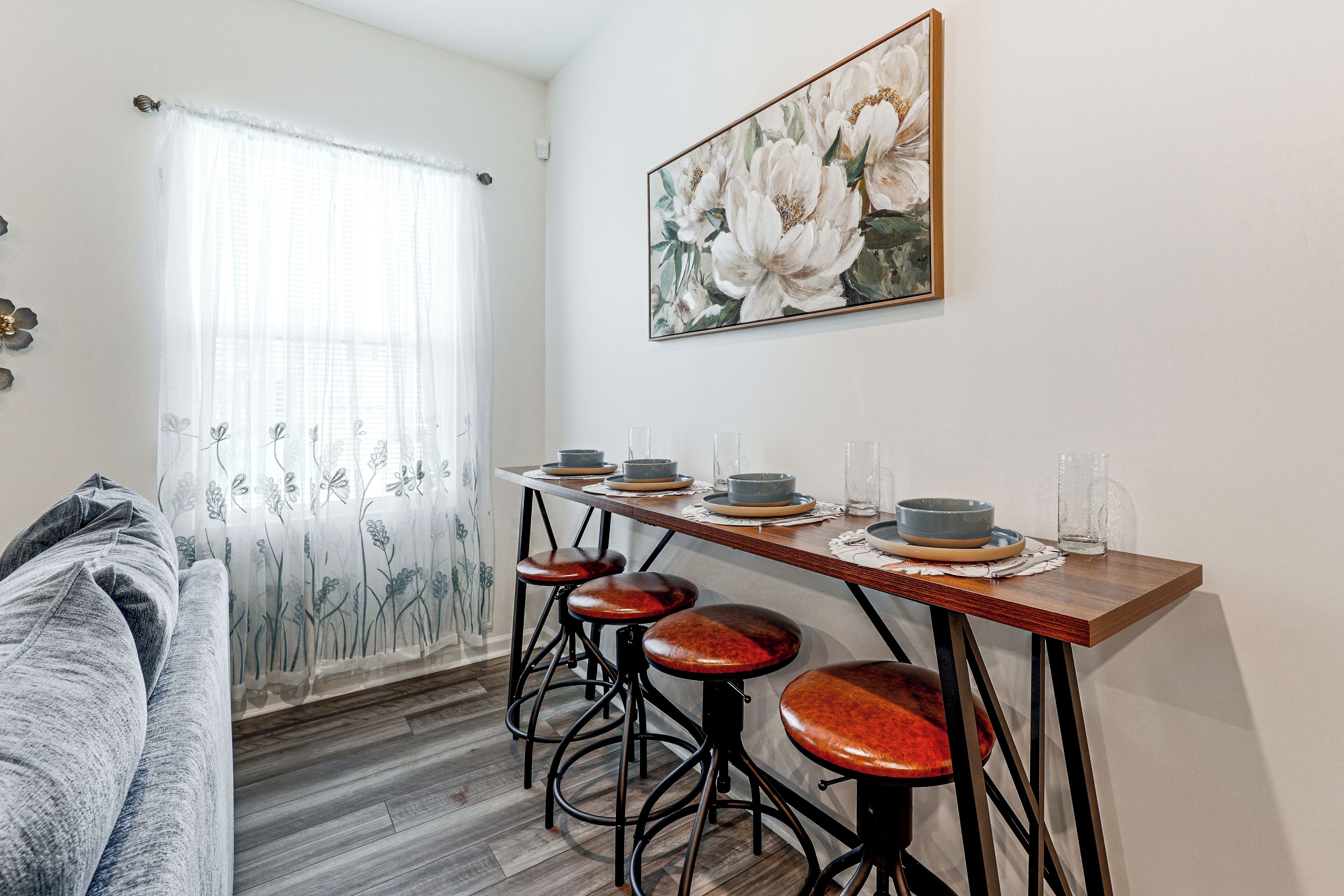 Un comedor con una mesa de madera, cuatro taburetes y un cuadro de flores en la pared.