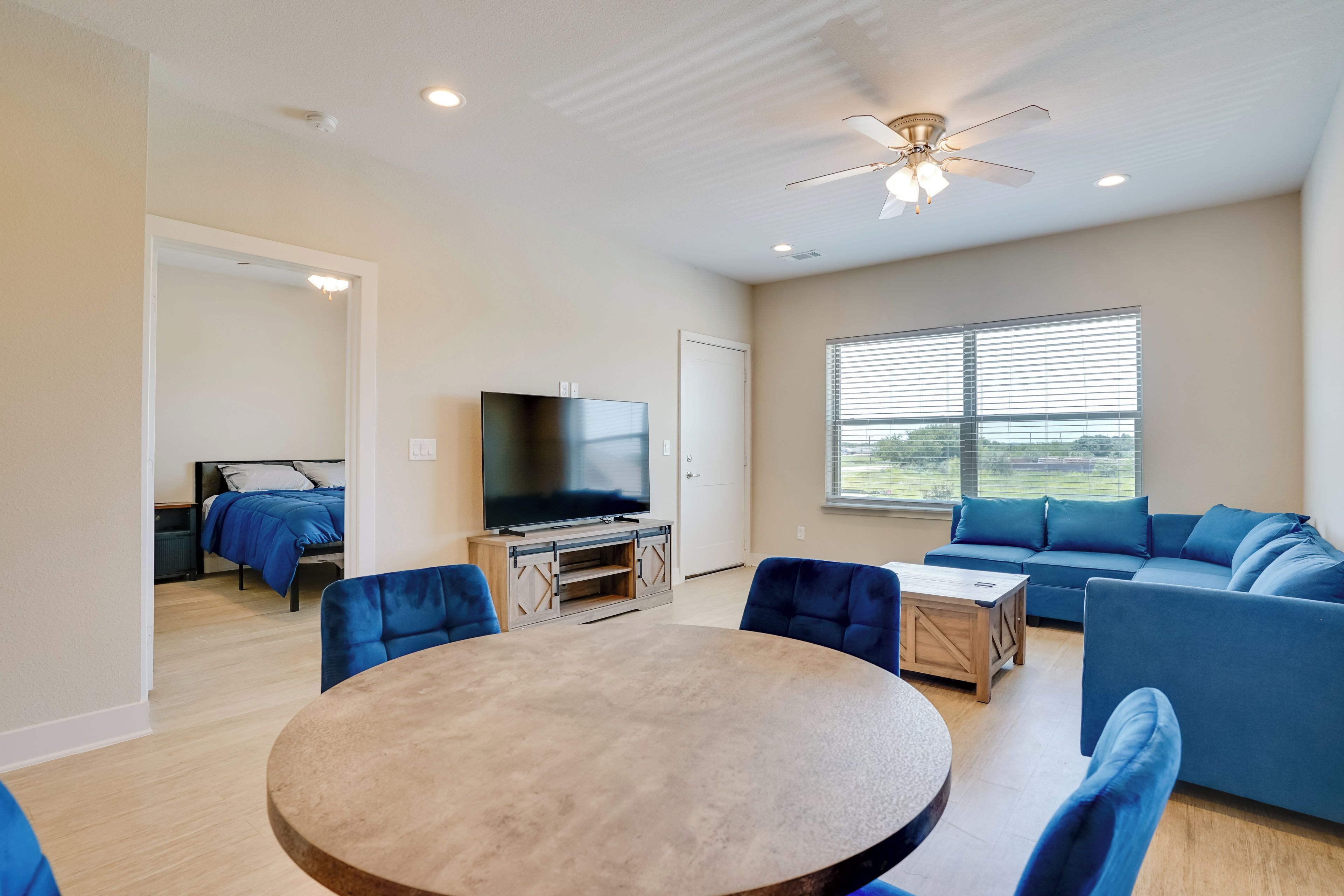 A spacious living room with a round table, blue sofas, a flat-screen TV, and a ceiling fan.
