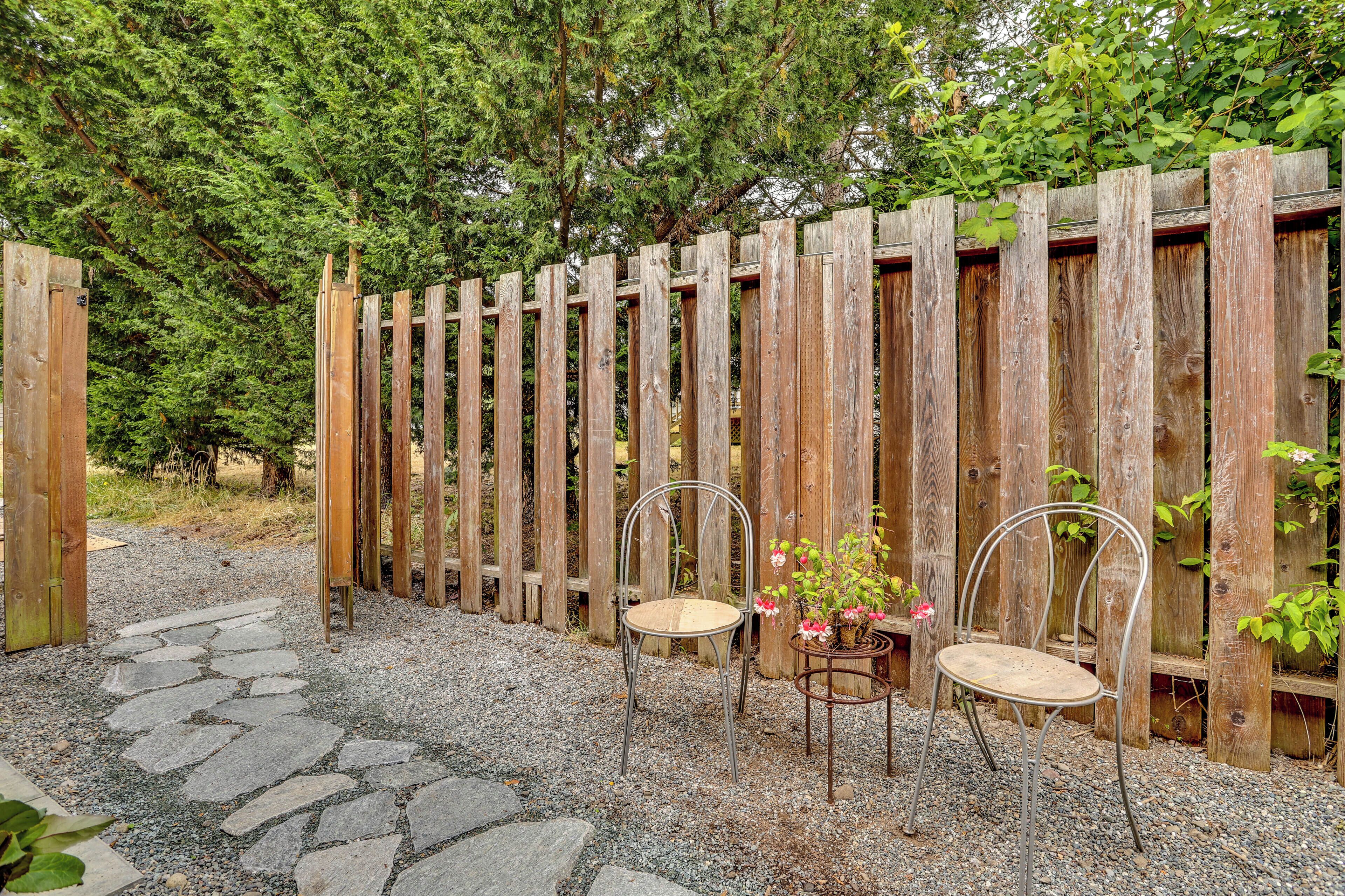 A wooden fence with two metal chairs and a table in front, surrounded by gravel and greenery.