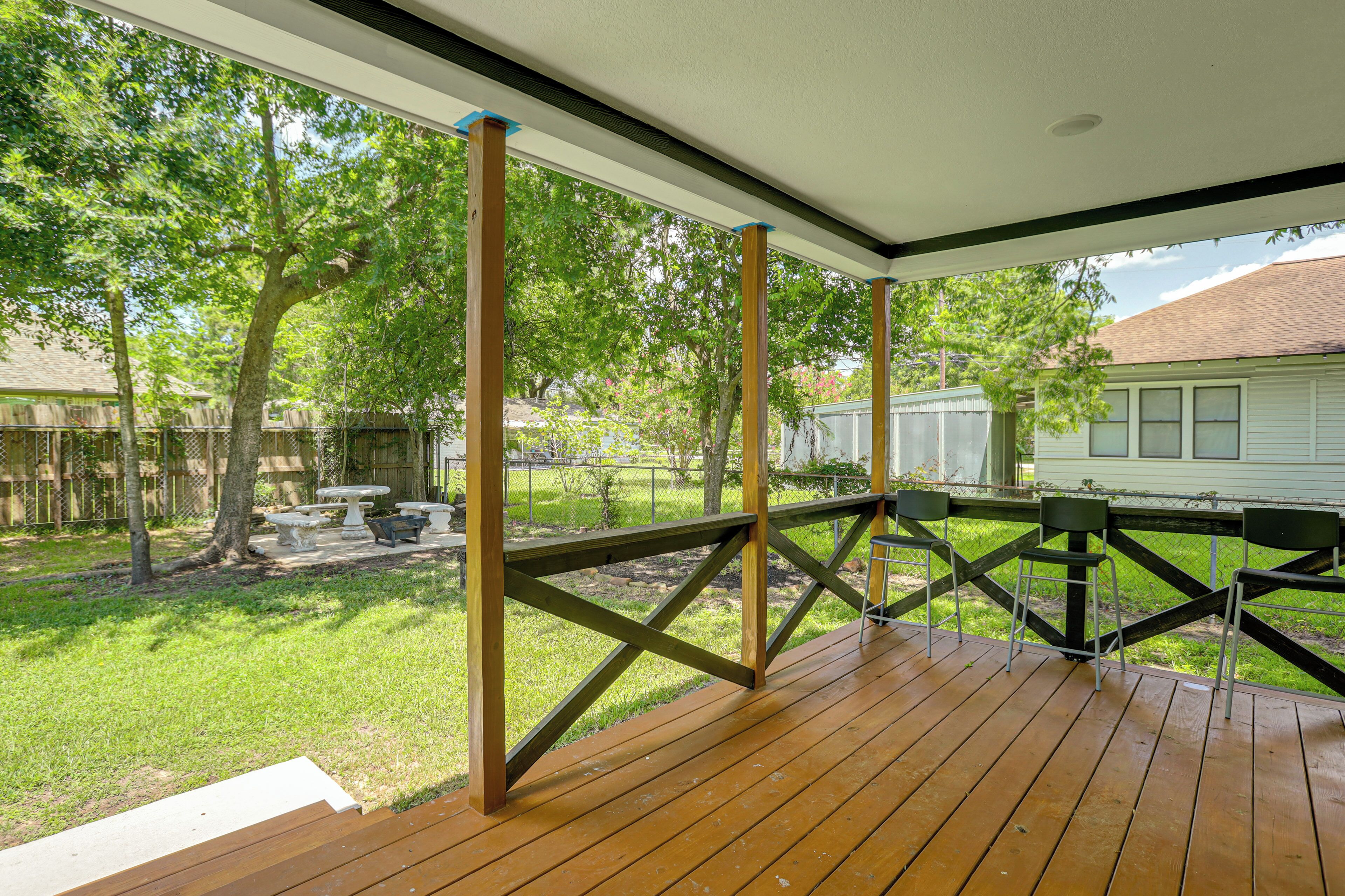 Une terrasse extérieure couverte, dotée d’un plancher en bois et entourée d’une rambarde en métal, offre une vue sur une cour verdoyante avec des arbres et une maison en arrière-plan.
