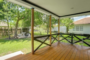Um deck coberto ao ar livre com piso de madeira, rodeado por um corrimão de metal, com vista para um gramado com árvores e uma casa ao fundo.