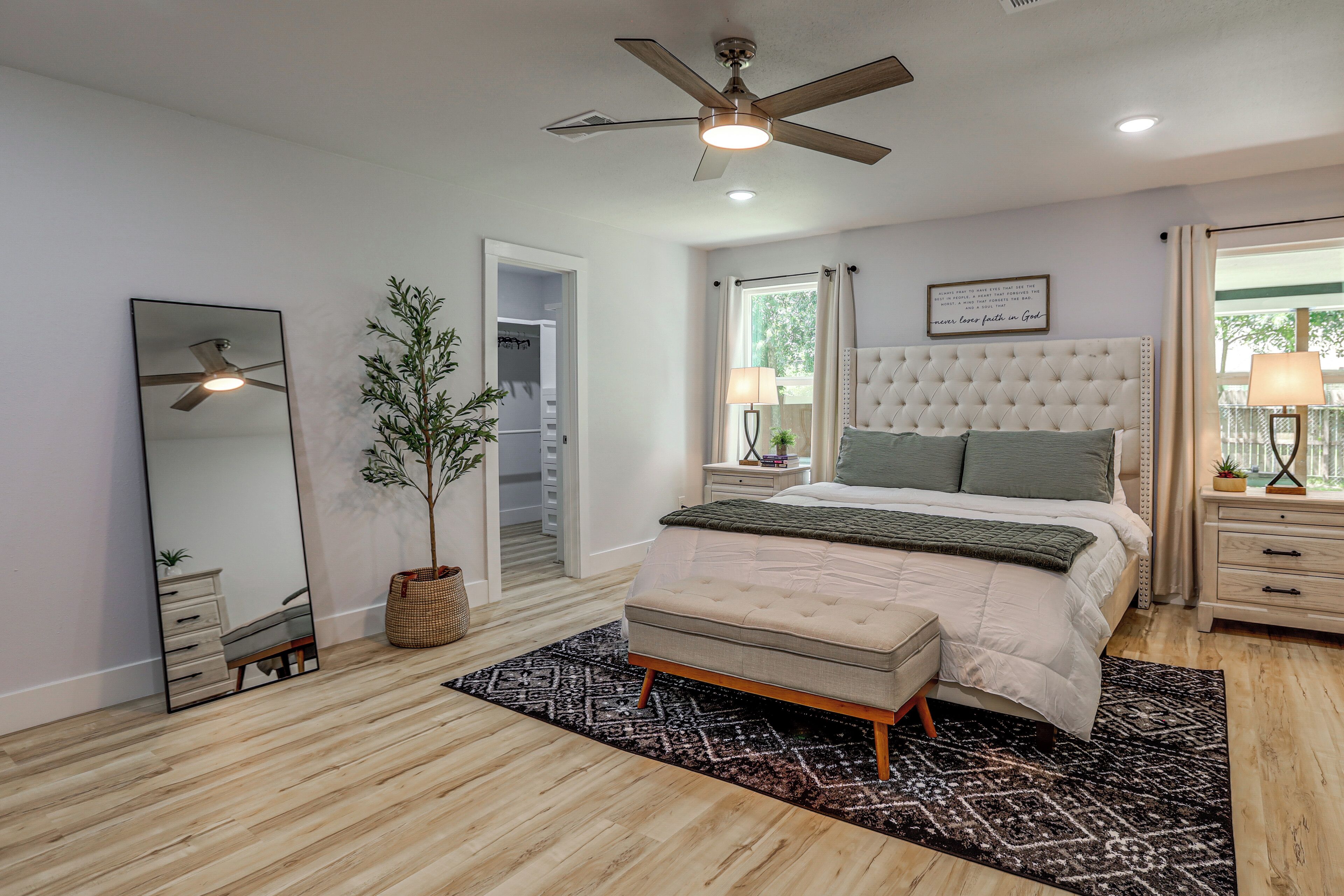 A bedroom with a bed, bedside tables, a ceiling fan, a mirror, a potted plant, and a window with curtains.