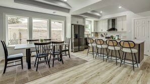 A modern kitchen with a dining area, featuring a large island with bar stools, a refrigerator, and a view through the windows. - Walk to Main St Tomball! Family Retreat w/ Yard (Tomball)