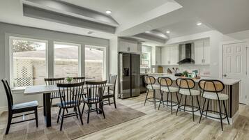 A modern kitchen with a dining area, featuring a large island with bar stools, a refrigerator, and a view through the windows.