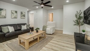 A modern living room with a black leather sofa, a wooden coffee table, a white armchair, and a ceiling fan. - Walk to Main St Tomball! Family Retreat w/ Yard (Tomball)