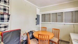Ein Zimmer mit einem runden Holztisch und zwei Stühlen, einem Fenster mit Jalousien, einer Wandsteckdose und einem karierten Kissen an der Wand.