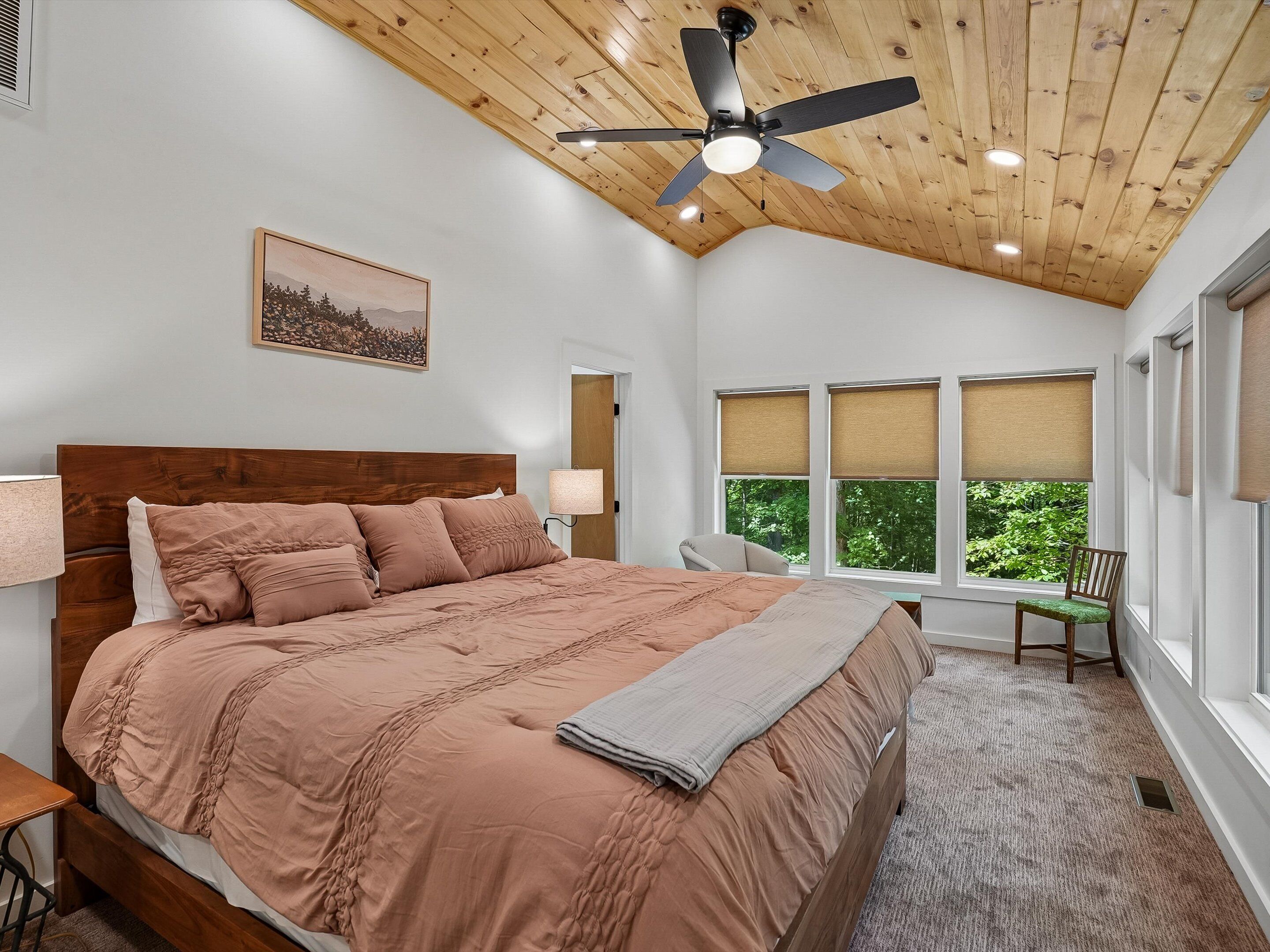 Un dormitorio con techo de madera, una cama con cabecera, una mesita de noche, una silla, una ventana con persianas y un ventilador de techo.