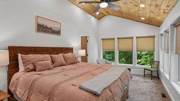 Un dormitorio con techo de madera, una cama con cabecera, una mesita de noche, una silla, una ventana con persianas y un ventilador de techo.