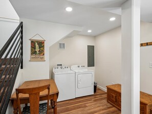 Un cuarto de lavandería con una mesa de madera, dos lavadoras, un basurero y un panel eléctrico instalado en la pared.
