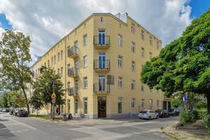 Un immeuble jaune à plusieurs étages, doté de balcons, avec une zone piétonne et des voitures garées.