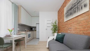 A modern kitchen with white cabinets, a dining table with chairs, and a grey sofa in a living area with a brick wall and a framed picture. - Stylish Apartment for 4 People | Near the Theatre (Warszawa)