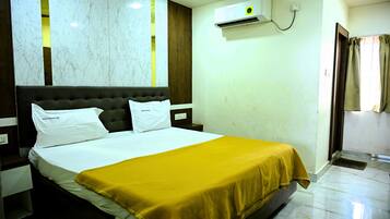 A hotel room with a bed, a nightstand, an air conditioning unit, and a window with curtains.