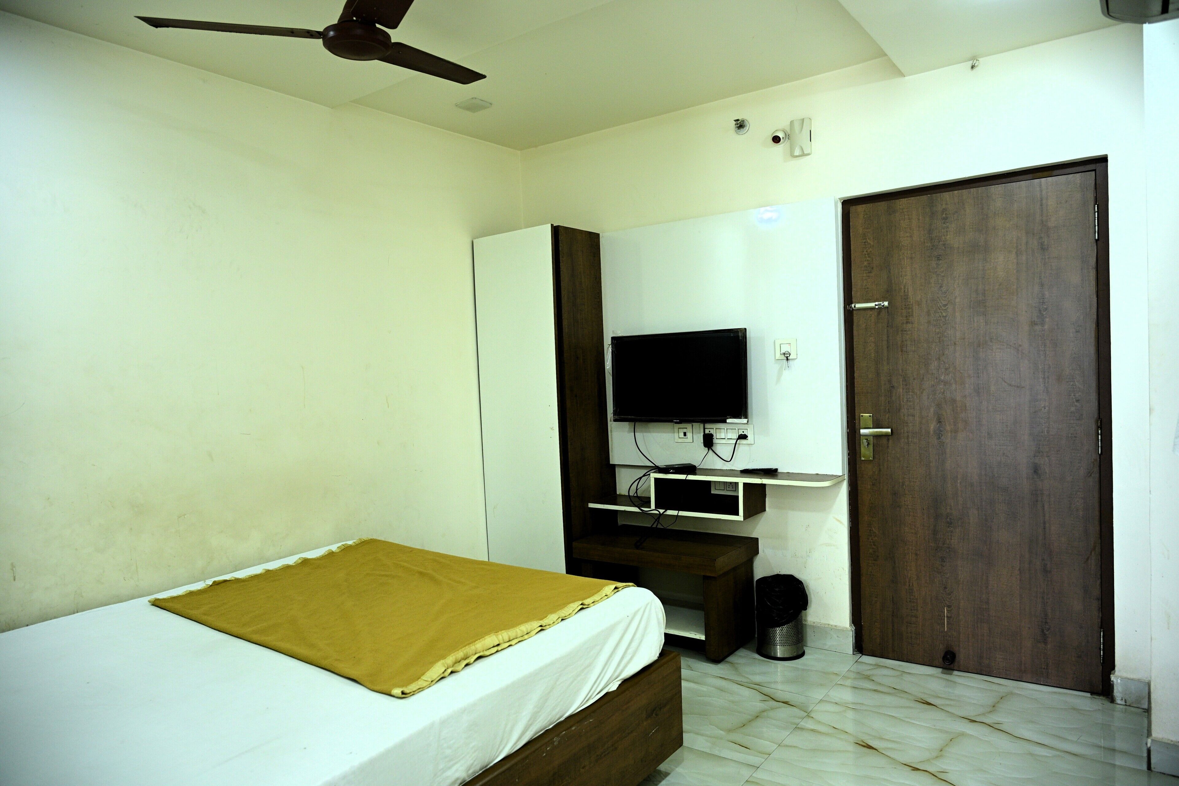 A bedroom with a bed, a television mounted on the wall, a ceiling fan, a wooden door, and a white wall.