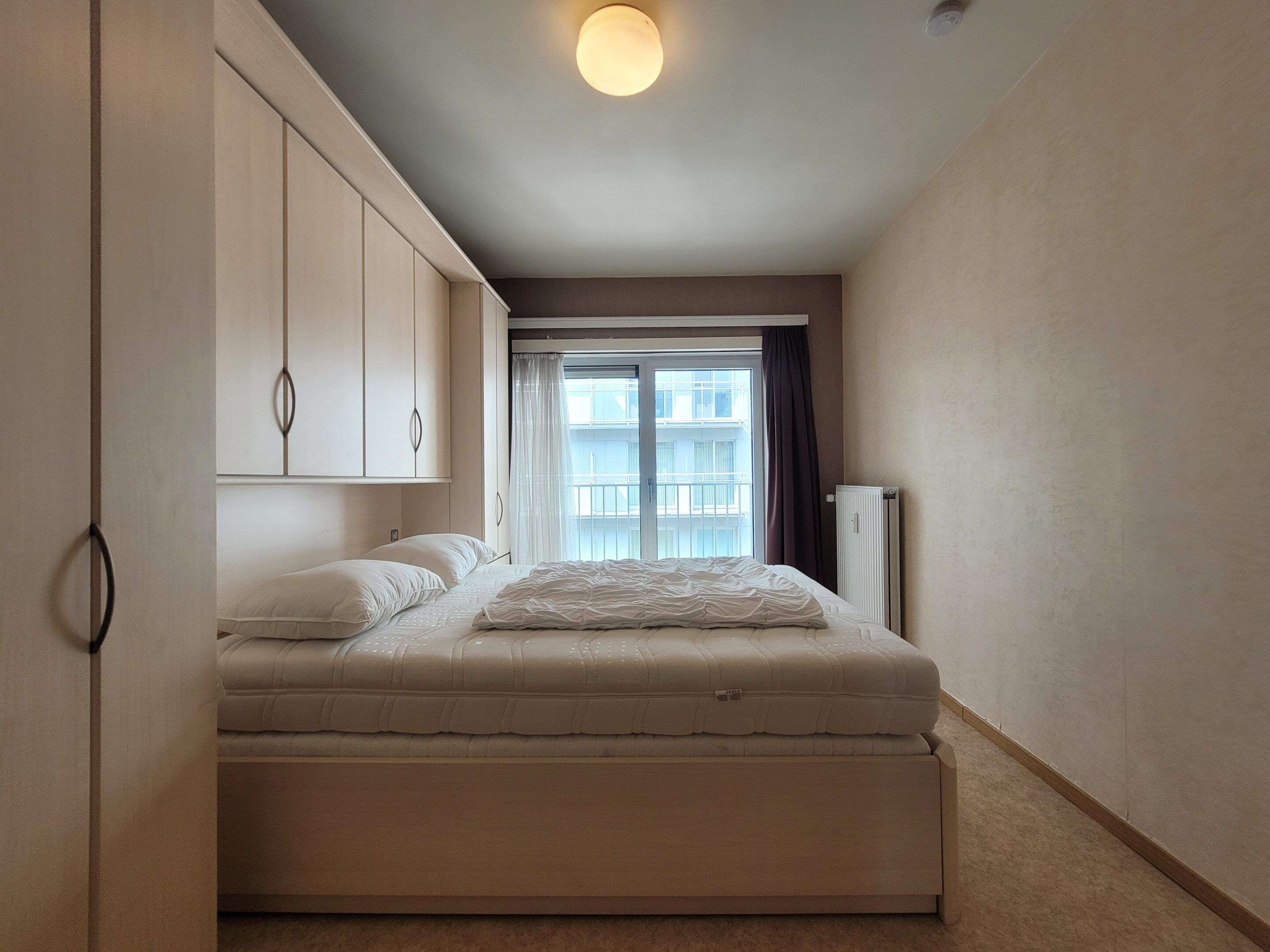 Een slaapkamer met een bed, een kledingkast, een radiator en een raam met gordijnen.