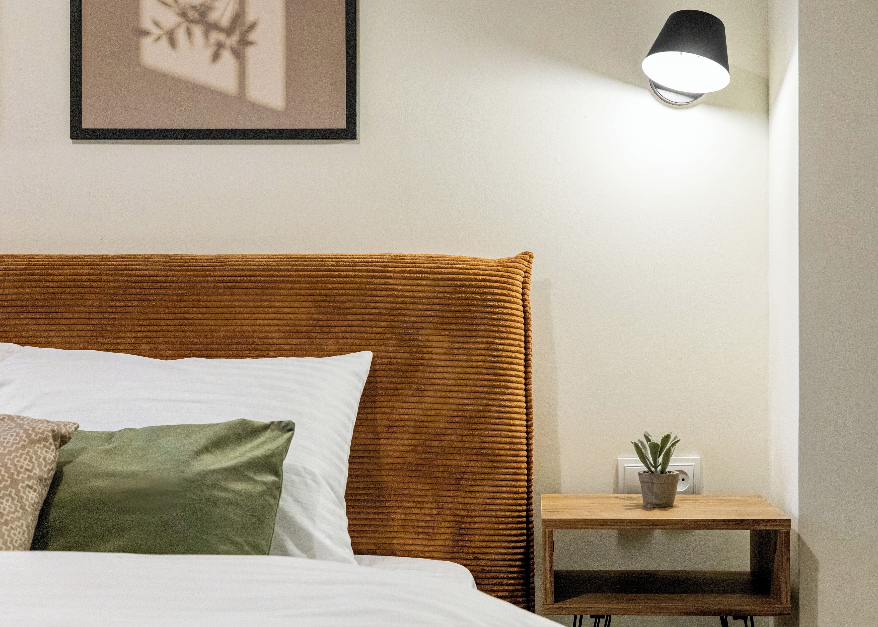 A neatly made bed with a wooden headboard, a bedside table with a potted plant, and a wall-mounted light fixture.