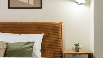 A neatly made bed with a wooden headboard, a bedside table with a potted plant, and a wall-mounted light fixture.