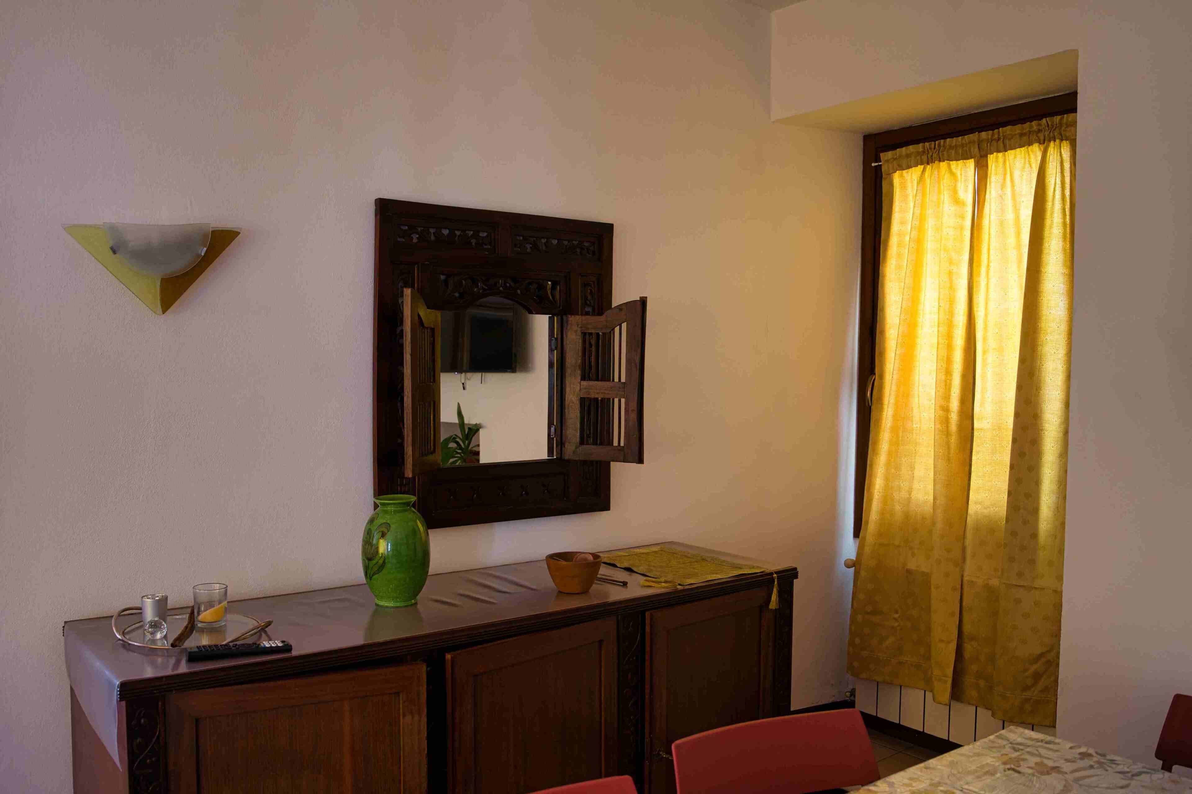 A room with a wooden desk, a mirror, a green vase, and a window with yellow curtains.