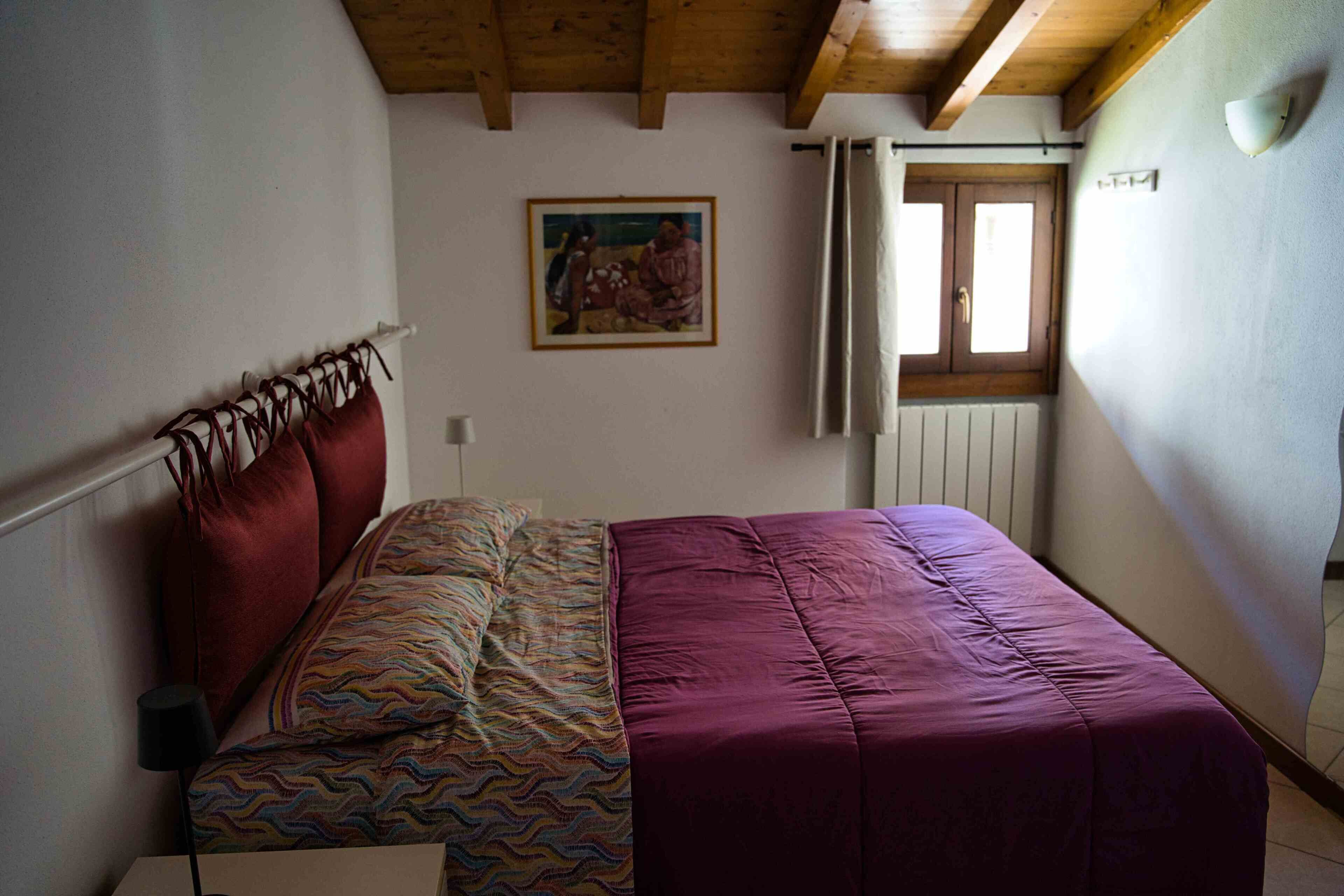 Un dormitorio con cama, un cuadro en la pared, una ventana con cortinas y un radiador.