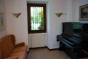 Un salón con piano, un radiador, una ventana que da a zonas verdes y un sofá marrón.