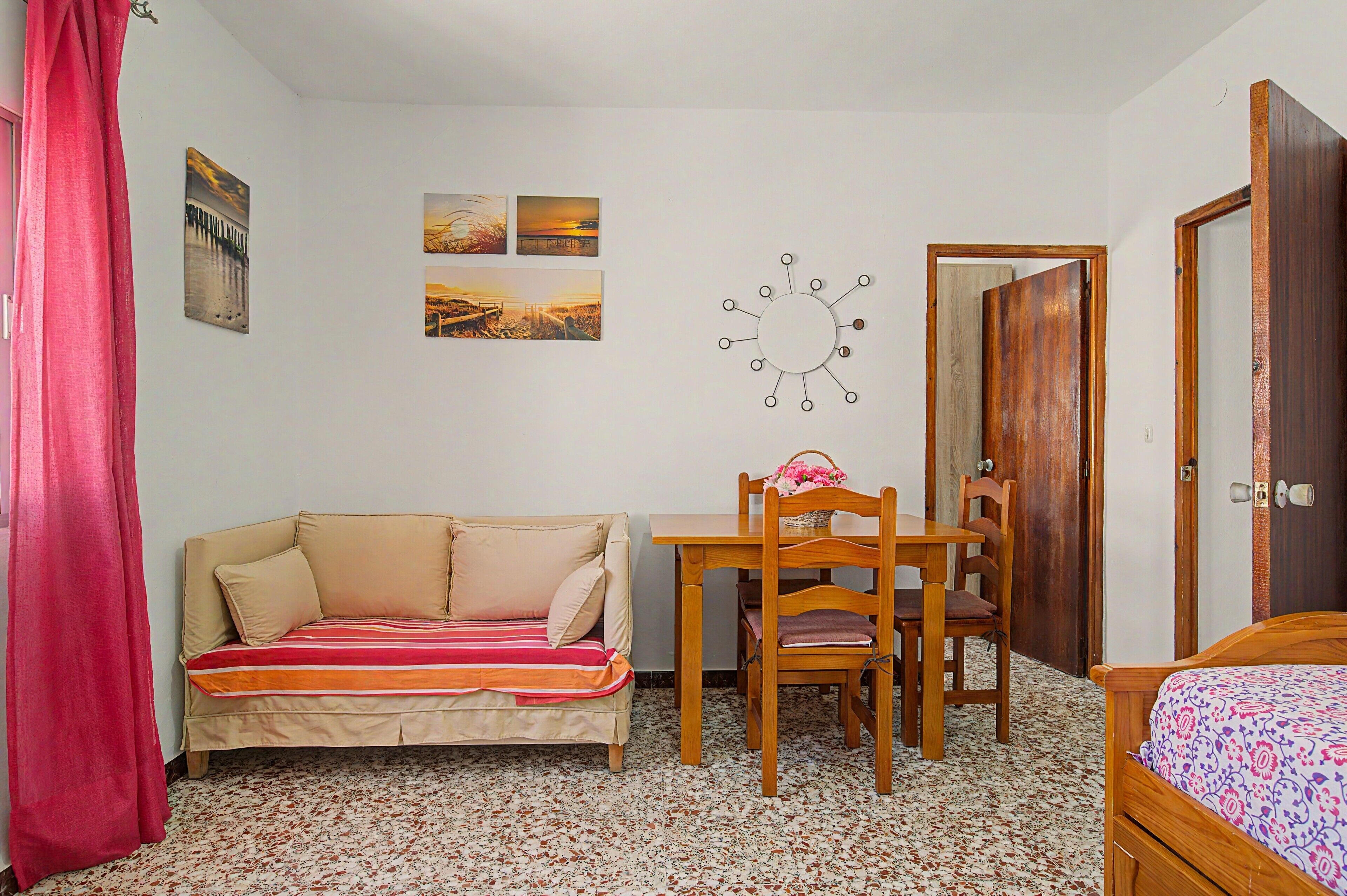 A small, furnished room with a sofa, a wooden table with chairs, a wall-mounted clock, and a door.