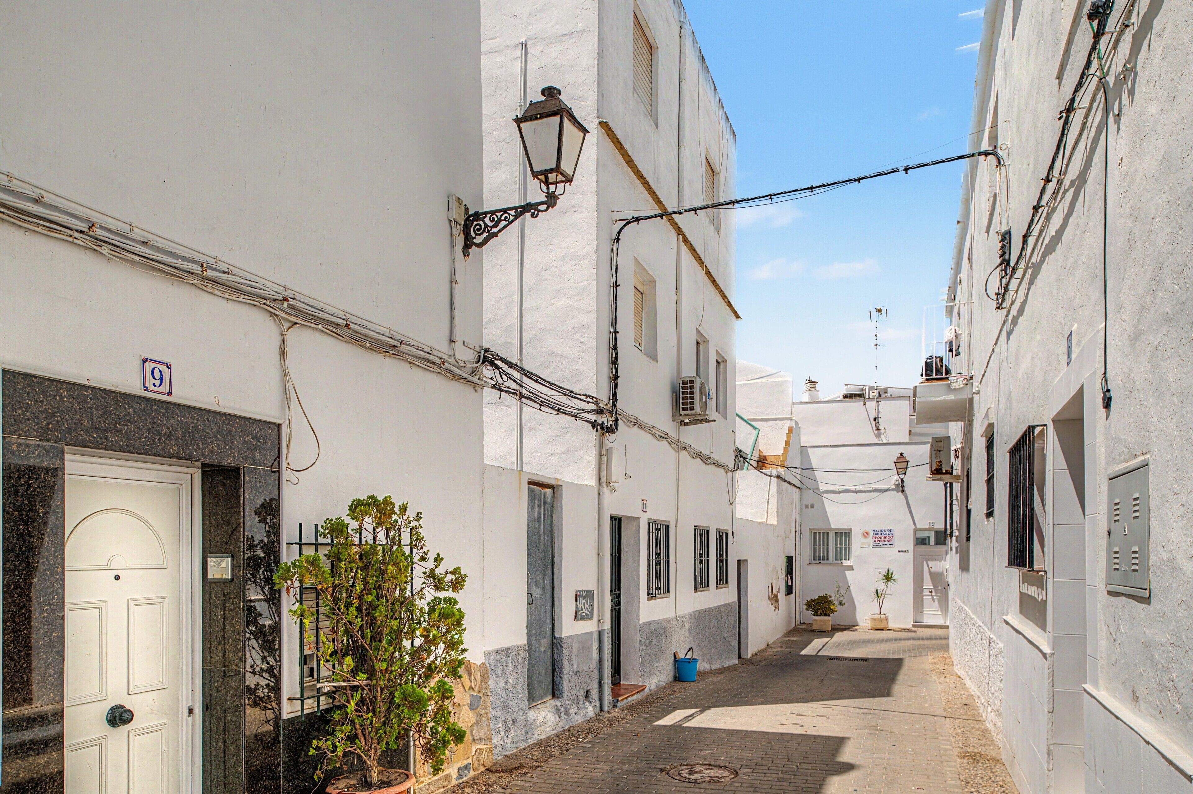 A narrow street with white buildings, a door with a number plate, and a small tree.