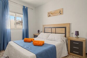 A neatly made bed with blue and white bedding, two lamps on nightstands, a painting above the bed, and a window with blue curtains. - El Rincón de Conil Apartment with Sea Views and Shared Terrace (Conil de la Frontera)