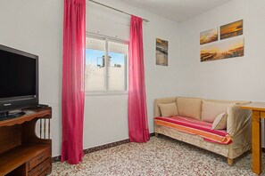 Ein Zimmer mit Fernseher auf einem Holzboden, einer Couch mit gestreiftem Überwurf, einem Fenster mit Vorhängen und Wandbildern.