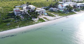 An aerial view of a coastal property with multiple buildings, a beach, and a person paddleboarding. - Villa Kakashki | Beachfront, Pool, and Padel (Yucatán)