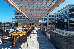 An outdoor patio with wooden tables and wicker chairs, a stone wall, and a grill area. - Refined Coastal Condo with Pool near Beach (Rehoboth Beach)