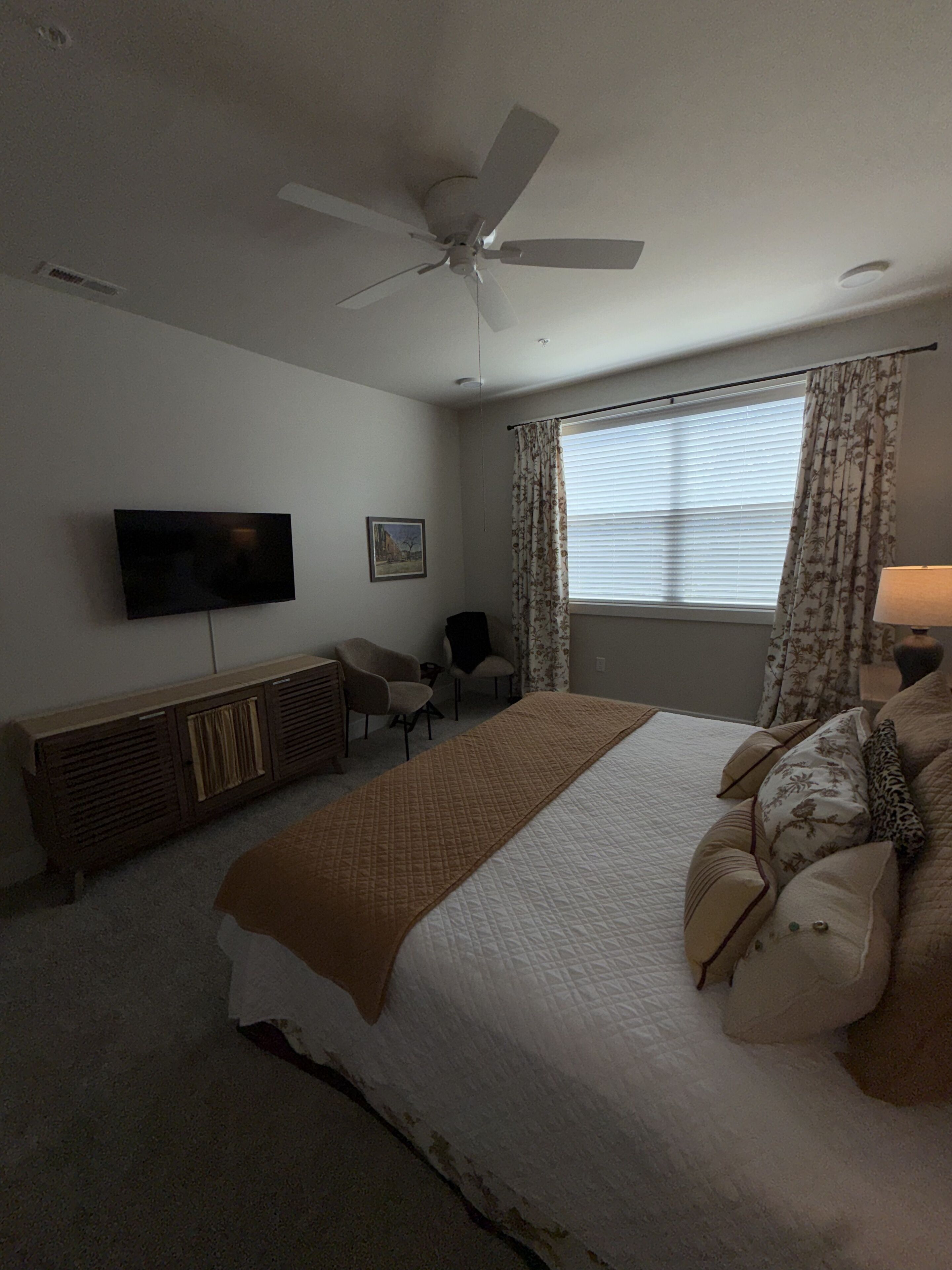 A bedroom with a bed, a television, a ceiling fan, a window with curtains, and a bedside table.