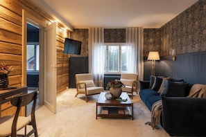 A cozy living room with a sofa, two armchairs, a coffee table with a vase, a flat-screen TV, and a window with curtains.