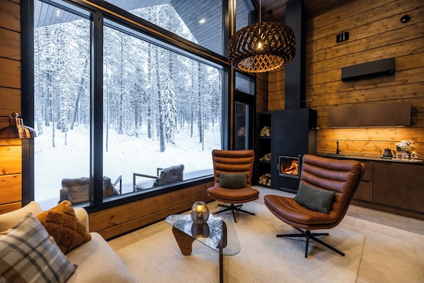 A cozy interior with a large window overlooking a snowy forest, a fireplace, leather armchairs, and a modern glass coffee table.
