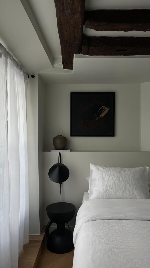 A bedroom with a bed, a bedside table, a lamp, a painting, and a window with curtains. - Studio Design à Palais Royal (Paris)
