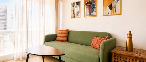 A living room with a green sofa, a wooden coffee table, and a side table with a lamp. There are abstract paintings on the wall and a window with curtains.