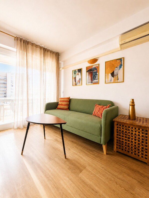 Een woonkamer met een groene bank, een houten salontafel en een bijzettafeltje met een lamp. Op de muur hangen abstracte schilderijen en er is een raam met gordijnen.
