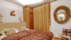A bedroom with a large bed, a wooden wardrobe, a small table, a mirror, and a lamp.