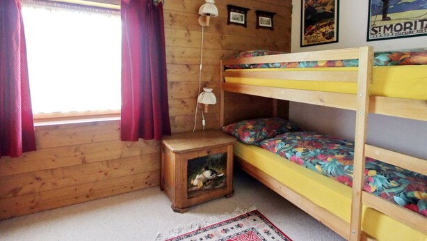 Ein Zimmer in einer Holzhütte mit Etagenbetten, einem kleinen Tisch mit Lampe und einem Fenster mit Vorhängen.