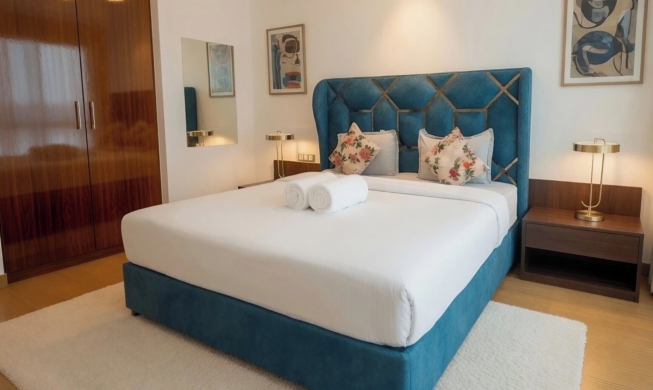 Una cama bien hecha con un cabecero azul, dos almohadas y toallas enrolladas. Hay dos mesitas de noche con lámparas y un armario de madera al fondo.