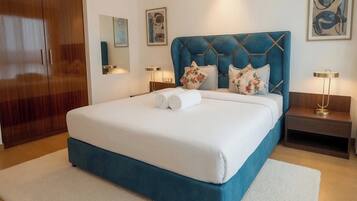 Una cama bien hecha con un cabecero azul, dos almohadas y toallas enrolladas. Hay dos mesitas de noche con lámparas y un armario de madera al fondo.