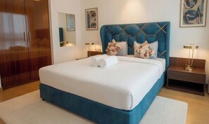 Una cama bien hecha con un cabecero azul, dos almohadas y toallas enrolladas. Hay dos mesitas de noche con lámparas y un armario de madera al fondo.
