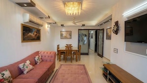 A living room with a sofa, a dining table with chairs, a television, and artwork on the walls. - Fortune Home Service Apartment E120 3BHK (Delhi)