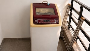 A compact, white and maroon laundry machine with a control panel, located on a tiled floor next to a staircase with a metal railing. - Fortune Home Service Apartment E120 3BHK (Delhi)
